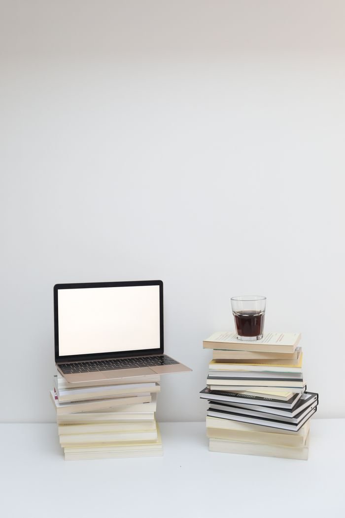 Opened laptop with empty screen and glass of fresh coffee placed on stacks of books on white table against white background